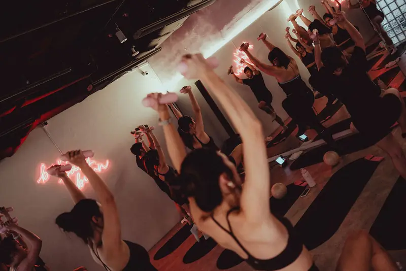 A hot mat class with arms raised, seen from above under pink neon